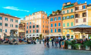 Une place animée à Rome, avec des bâtiments colorés et une fontaine. Un panneau indique "Take Away Cocktail 4.50", un clin d'œil au "saint amour menu".