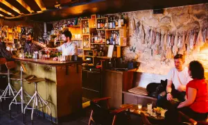 Ambiance feutrée dans ce bar caché. Un barman sert un cocktail, des clients discutent au comptoir et sur une banquette.