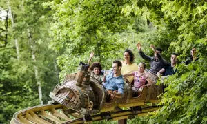 Famille souriante et joyeuse sur un wagon de parc attraction, bras levés, profitant d'une descente dans un décor verdoyant.