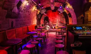 Scène de concert dans le caveau des oubliettes, éclairée par des lumières violettes et rouges. Tables et tabourets disposés devant la scène où se trouve une batterie et des guitares.