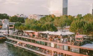 Un bateau-mouche animé sur la Seine, avec des gens faisant la fête sur le pont. Le soleil brille sur un gratte-ciel, créant un effet de "mazette" lumineux.