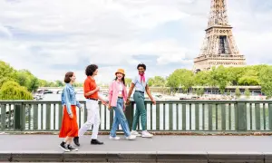 Quatre jeunes amis profitent d'une promenade ensoleillée à Paris, avec la Tour Eiffel en arrière-plan. L'un d'eux, un étudiant, porte un foulard rose vif.