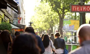 Des passants se promènent sur un grand boulevard parisien, sous un soleil radieux. Un panneau "METRO" rouge indique une station.