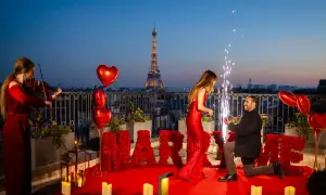 Proposition romantique à Paris, avec la Tour Eiffel en arrière-plan. Un homme s'agenouille, une bague à la main, devant sa compagne. Des roses rouges forment le mot "MARRY ME". Une violoniste joue, des feux d'artifice illuminent la scène. Un moment dig...
