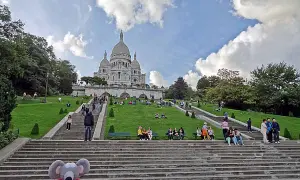 Un adorable raton laveur en pull rouge pose sur les marches menant au Sacré-Cœur, où des visiteurs profitent du soleil sur l'herbe verdoyante.