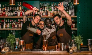 Quatre barmans joyeux préparent des cocktails au bar "Le Renard". L'un verse un liquide d'une bouteille rouge, un autre d'une bouteille transparente.