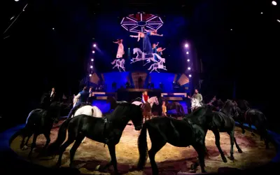 Spectacle équestre des folies Gruss : des chevaux noirs et blancs évoluent sous les projecteurs, tandis que des acrobates aériennes dansent au-dessus.