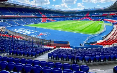 Vue panoramique du **Parc des Princes** vide, avec ses tribunes bleues et rouges, son terrain de football verdoyant et le logo du PSG au centre.