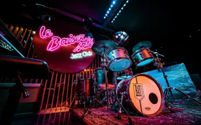 Batterie de jazz dans un bar parisien, éclairé par des néons roses et bleus. Ambiance feutrée pour une soirée musicale.