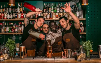 Quatre barmans joyeux préparent des cocktails au bar "Le Renard". L'un verse un liquide d'une bouteille rouge, un autre d'une bouteille transparente.