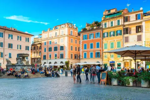 Une place animée à Rome, avec des bâtiments colorés et une fontaine. Un panneau indique "Take Away Cocktail 4.50", un clin d'œil au "saint amour menu".