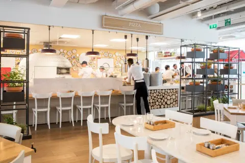 Un restaurant italien moderne avec un four à pizza visible. Des chefs préparent des plats derrière le comptoir, tandis que des tables sont dressées pour les clients.