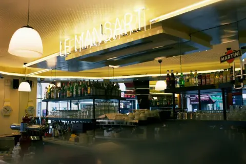 Bar lumineux avec enseigne néon "LE MANSART PARIS". Bouteilles d'alcool et verres alignés sur les étagères.