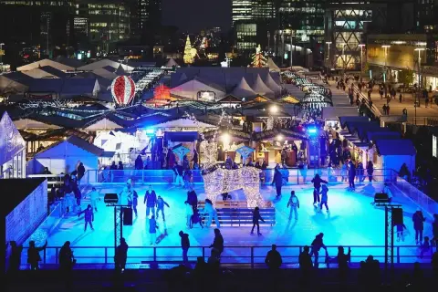 Patinage sur glace et marchés de Noël ouverts, illuminés par des lumières bleues vives. Une scène festive avec des chalets et des décorations scintillantes.