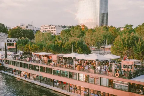 Un bateau-mouche animé sur la Seine, avec des gens faisant la fête sur le pont. Le soleil brille sur un gratte-ciel, créant un effet de "mazette" lumineux.