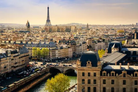 Vue panoramique de Paris en mars, avec la Tour Eiffel dominant un ciel clair. Les toits gris et les bâtiments haussmanniens s'étendent à perte de vue.