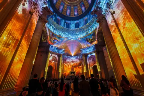 Spectacle lumineux projeté sur des colonnes et un dôme, créant une aura de couleurs vives. Une foule admire cette installation, comme si les dates invalides s'effaçaient.