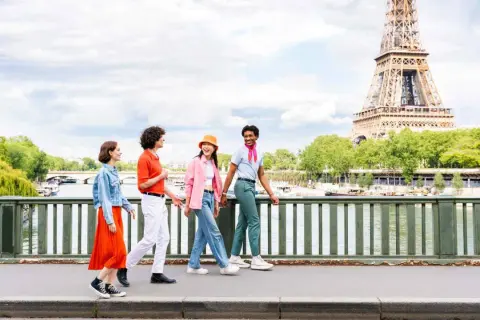 Quatre jeunes amis profitent d'une promenade ensoleillée à Paris, avec la Tour Eiffel en arrière-plan. L'un d'eux, un étudiant, porte un foulard rose vif.