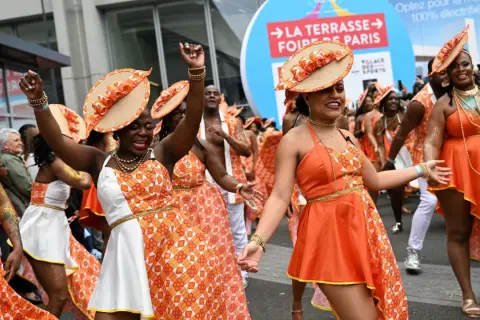 Des danseuses en tenues orange et blanches, chapeaux assortis, célèbrent la foire de Paris 2026 dans une ambiance festive.