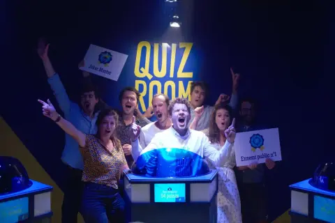 Équipe joyeuse dans une "Quiz Room" à Paris La Villette, célébrant une victoire. Ils tiennent des pancartes "Joker Master" et "Ennemi public n°1".