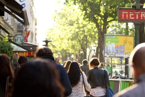 Des passants se promènent sur un grand boulevard parisien, sous un soleil radieux. Un panneau "METRO" rouge indique une station.