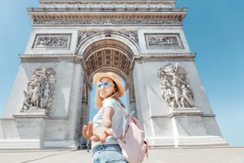 Une jeune femme tend la main devant l'Arc de Triomphe. L'accès au monument est gratuit, une belle opportunité de visite.