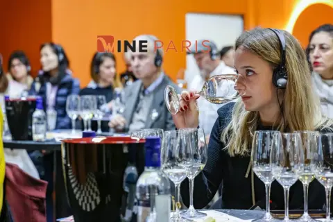 Dégustation de vin à Wine Paris 2026. Une femme hume un verre, casque sur les oreilles, entourée d'autres participants.