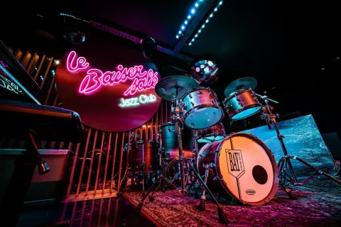 Batterie de jazz dans un bar parisien, éclairé par des néons roses et bleus. Ambiance feutrée pour une soirée musicale.