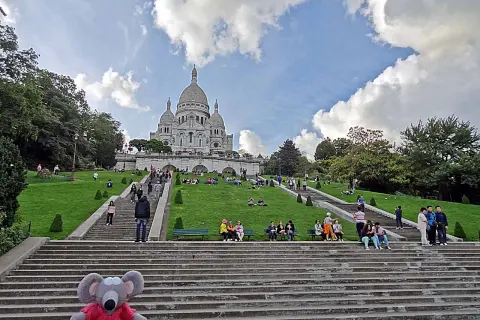 Un adorable raton laveur en pull rouge pose sur les marches menant au Sacré-Cœur, où des visiteurs profitent du soleil sur l'herbe verdoyante.