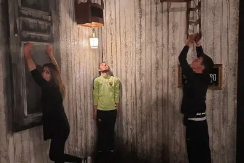 Trois personnes dans une pièce aux murs décrépits, défiant la gravité. Une femme s'accroche à un mur, un homme flotte, un autre escalade une chaise. Ambiance de **the dark dreams Paris**.