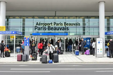Voyageurs avec bagages devant l'aéroport Paris-Beauvais, prêts pour leur vol.