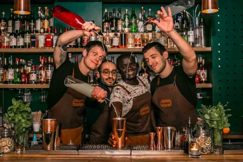 Quatre barmans joyeux préparent des cocktails au bar "Le Renard". L'un verse un liquide d'une bouteille rouge, un autre d'une bouteille transparente.