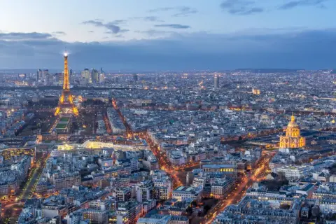 Vue panoramique de Paris au crépuscule, avec la Tour Eiffel illuminée et le dôme des Invalides. On aperçoit le quartier de La Défense au loin. Un panorama depuis l'**étage tour Montparnasse**.