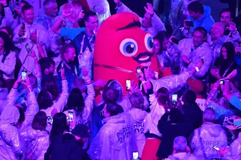 La Phryge JO, mascotte des JO, interagit avec une foule joyeuse. Les gens filment et prennent des photos avec leurs téléphones.