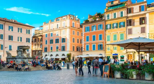 Une place animée à Rome, avec des bâtiments colorés et une fontaine. Un panneau indique "Take Away Cocktail 4.50", un clin d'œil au "saint amour menu".