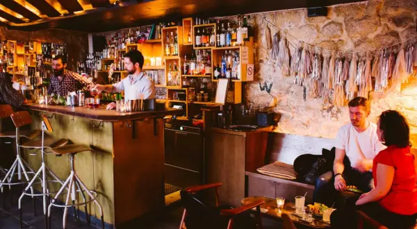 Ambiance feutrée dans ce bar caché. Un barman sert un cocktail, des clients discutent au comptoir et sur une banquette.