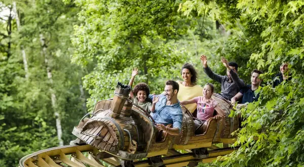 Famille souriante et joyeuse sur un wagon de parc attraction, bras levés, profitant d'une descente dans un décor verdoyant.
