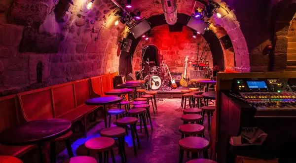 Scène de concert dans le caveau des oubliettes, éclairée par des lumières violettes et rouges. Tables et tabourets disposés devant la scène où se trouve une batterie et des guitares.
