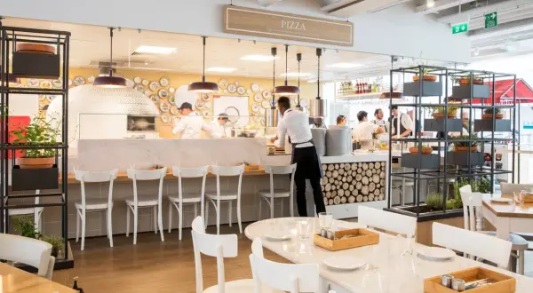 Un restaurant italien moderne avec un four à pizza visible. Des chefs préparent des plats derrière le comptoir, tandis que des tables sont dressées pour les clients.