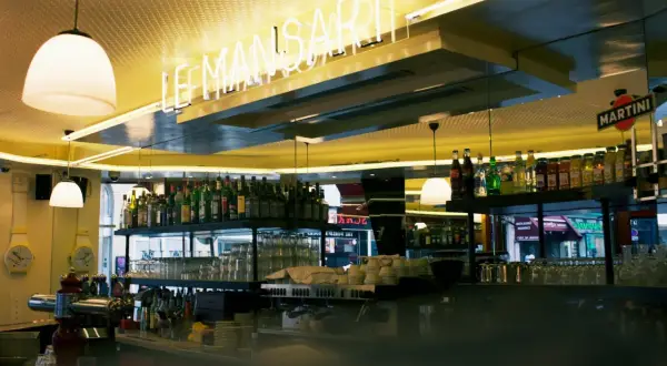 Bar lumineux avec enseigne néon "LE MANSART PARIS". Bouteilles d'alcool et verres alignés sur les étagères.