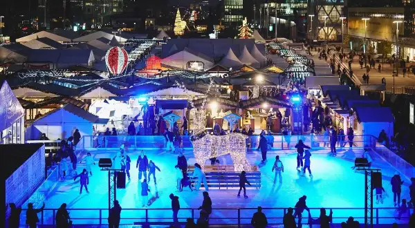Patinage sur glace et marchés de Noël ouverts, illuminés par des lumières bleues vives. Une scène festive avec des chalets et des décorations scintillantes.