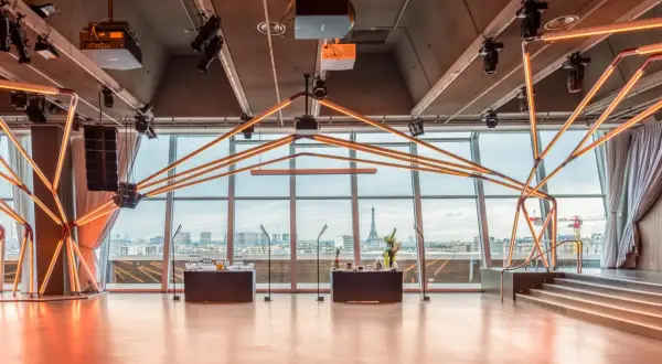 Salle de réception moderne avec vue panoramique sur Paris, éclairage néon, bar et scène. La Tour Eiffel est visible au loin.