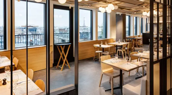 Restaurant moderne avec vue sur Paris. Tables dressées, lumières douces, et le logo "Printemps du Goût" visible.