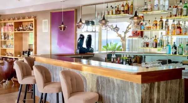 Un **majestic bar** avec des tabourets beiges, des étagères remplies de bouteilles et une vue sur la mer.