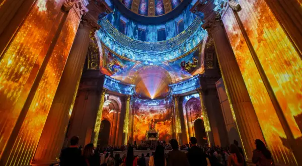 Spectacle lumineux projeté sur des colonnes et un dôme, créant une aura de couleurs vives. Une foule admire cette installation, comme si les dates invalides s'effaçaient.