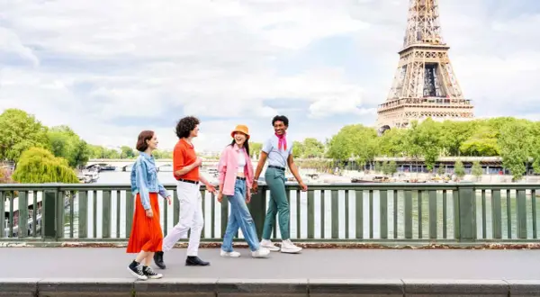 Quatre jeunes amis profitent d'une promenade ensoleillée à Paris, avec la Tour Eiffel en arrière-plan. L'un d'eux, un étudiant, porte un foulard rose vif.