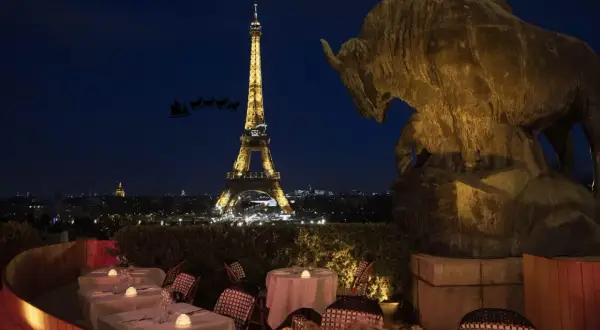 Vue nocturne de Paris avec la Tour Eiffel illuminée. Un bar ouvert le 25 décembre, avec des tables dressées et une statue de taureau. Le traîneau du Père Noël traverse le ciel.