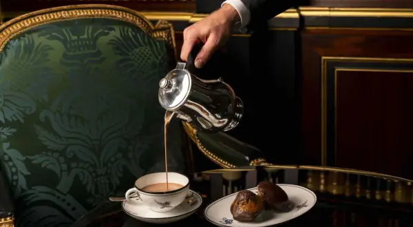 Un homme verse un chocolat chaud dans une tasse, accompagné de madeleines, dans un salon parisien luxueux.