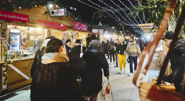 Ambiance féerique d'un marché de Noël à Paris. Des chalets en bois illuminés proposent des spécialités, tandis que des passants flânent sous les guirlandes lumineuses.