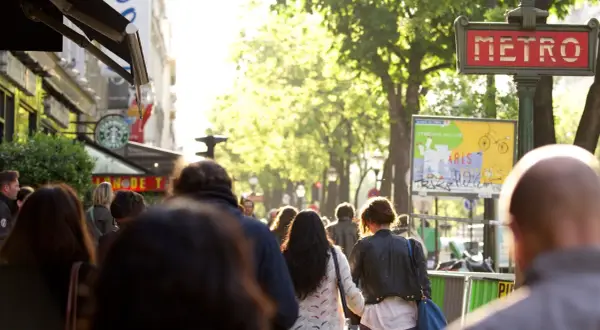 Des passants se promènent sur un grand boulevard parisien, sous un soleil radieux. Un panneau "METRO" rouge indique une station.